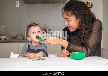 Junge Kleinkinder, die von einem lächelnden Hausmeister in einer gemütlichen Küche gefüttert werden. Stockfoto