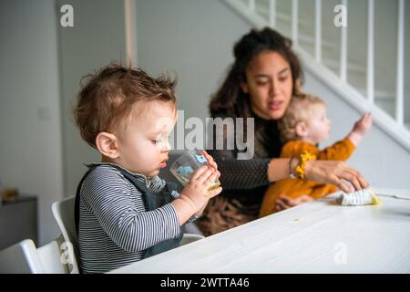 Ein kleines Kind schlürft aus einer Tasse, während eine Mutter am Esstisch über zwei Kinder wacht. Stockfoto