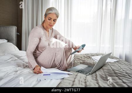 Frau, die am Laptop im Bett arbeitet, während sie das Telefon hält Stockfoto