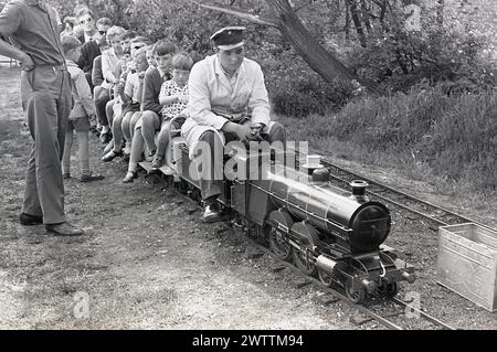 1960er Jahre, historische, sommerliche und externe Familien, die eine Fahrt mit einer Miniaturbahn genießen, Zugführer mit Hut, der die Miniatur-Dampfeisenbahn fährt, England, Großbritannien. Stockfoto