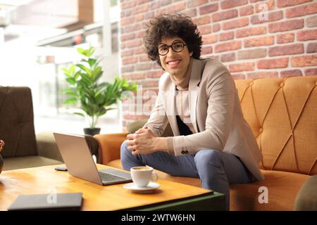 Ein lächelnder junger Mann in einem Café, der einen Kaffee trinkt und an einem Laptop arbeitet Stockfoto