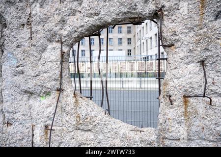 Dieses Foto zeigt ein Loch in einem verbleibenden Teil der Berliner Mauer aus nächster Nähe. Die verwitterte Textur und die freiliegende, rostfreie Bewehrung erzählen von Alter und Witterungseinwirkung. Durch das Loch ist ein gut erhaltenes Gebäude und ein Metallzaun zu sehen, der einen starken Kontrast zwischen Vergangenheit und Gegenwart darstellt Stockfoto