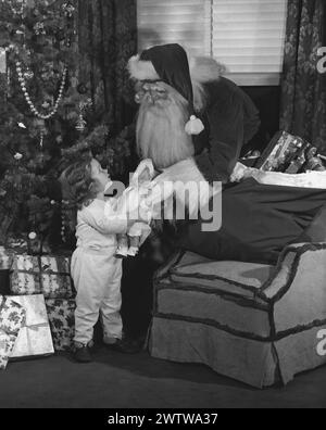 Der Weihnachtsmann steht zwischen dem Baum und einem Stuhl mit seiner Tasche mit Spielzeug auf dem Stuhl und überreicht einem kleinen Mädchen eine neue Puppe Stockfoto