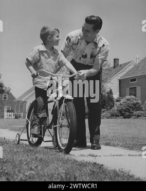 Dad und Sohn halten inne, um sich gegenseitig anzusehen, während sie lernen, Fahrrad auf dem Bürgersteig in einem Wohnviertel zu fahren. Stockfoto