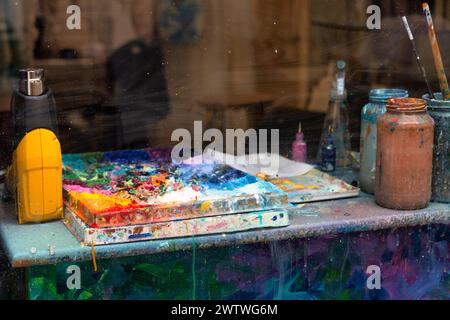 Ein Blick aus der Nähe auf den Arbeitsbereich eines Künstlers mit einer unordentlichen Farbpalette und fleckigen Gläsern, die mit Pinseln gefüllt sind Stockfoto