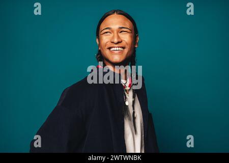Ein selbstbewusster, fröhlicher und stilvoller junger Mann mit geflochtenen Haaren steht vor einem blauen Hintergrund. Ethnischer Schmuck und legere Kleidung ergänzen sein einzigartiges Stockfoto