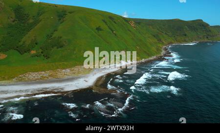 Wilde Natur, grüne Berge mit steilen grünen Hängen, die zum Meer führen. Clip. Die Luft aus Schaum blauer Wellen bricht in vulkanischem Gestein. Stockfoto