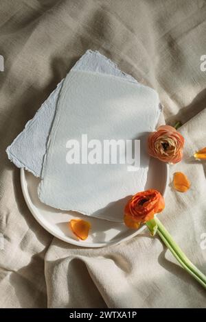 Blick über die Draufsicht auf leere handgefertigte Papierkarten auf einer Keramikplatte mit orangen Ranunkelblüten auf beiger Tischdecke Stockfoto