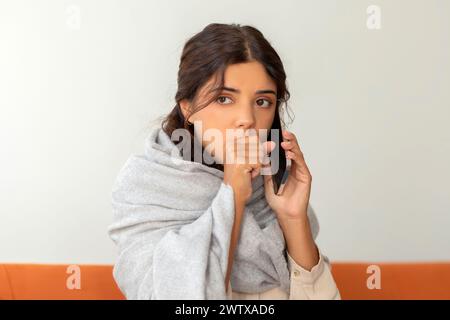 Hustende junge Frau, in Decke gehüllt, die zu Hause auf dem Handy spricht Stockfoto