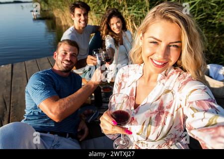 Gruppe von Freunden, die Spaß beim Picknick in der Nähe eines Sees haben, auf dem Pier sitzen und Wein trinken und Selfie machen. Stockfoto