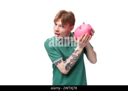 Fröhlich fröhlicher junger rothaariger Kerl in einem grünen T-Shirt hält eine Sparkasse mit Geld und denkt, wo er ausgeben soll Stockfoto