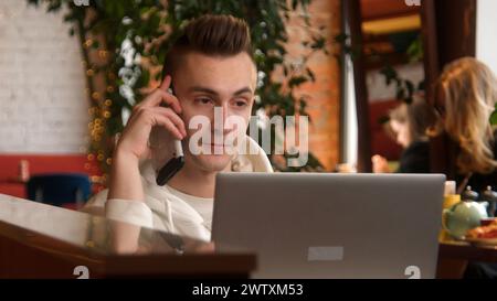 Der junge Mann telefoniert am Laptop im Café. Archivmaterial. Der junge Mann spricht am Telefon, während er am Laptop arbeitet. Der Mann nimmt Arbeitsanrufe entgegen Stockfoto