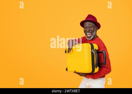 Fröhlicher älterer schwarzer Mann mit rotem Hut und Pullover, der einen hellgelben Koffer hält Stockfoto