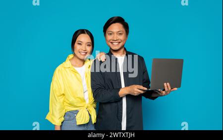 Ein lächelndes asiatisches Paar steht eng zusammen, der Mann hält einen Laptop und die Frau lehnt sich auf seine Schulter Stockfoto