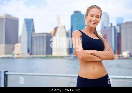 Frau und Lächeln am Fluss mit Blick auf die Stadt auf Sportkleidung zum Joggen, Fitness und gesunde Einstellung in New York. Porträt, Arme gefaltet und weibliche Person Stockfoto