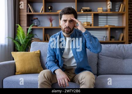 Ein nachdenklicher, erwachsener Mann mit Bart sitzt auf einem Sofa und wirkt gestresst oder gestört, umgeben von einer gemütlichen Wohnumgebung. Stockfoto
