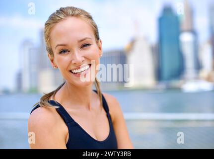 Porträt, Frau und Lächeln am Fluss mit Blick auf die Stadt auf Sportkleidung zum Joggen, Fitness und gesunde Einstellung in New York. Nahaufnahme, weibliche Person und Stockfoto