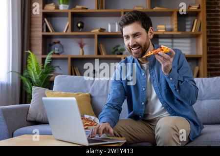 Glücklicher Mann in lässiger Kleidung, der ein Stück Pizza isst und an seinem Laptop arbeitet, auf einer Couch in einem gemütlichen Zuhause sitzt. Stockfoto