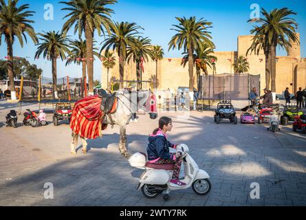 Ein junges Mädchen fährt mit einem Spielzeugroller an einem dekorierten Pferd auf dem Bourguiba-Platz, neben dem Ribat, Monastir, Tunesien Stockfoto