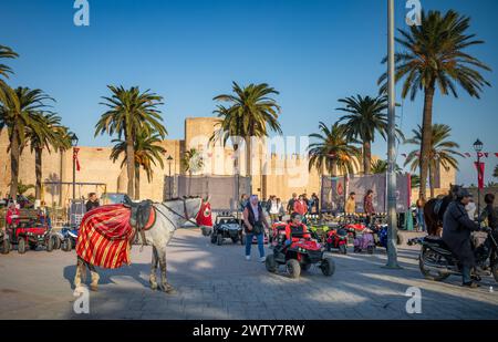 Ein junges Mädchen fährt mit einem Spielzeugroller an einem dekorierten Pferd auf dem Bourguiba-Platz, neben dem Ribat, Monastir, Tunesien Stockfoto