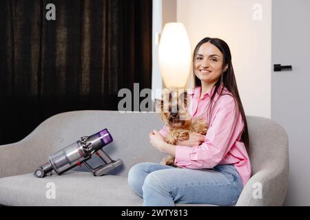 Eine süße junge Frau sitzt mit ihrem Hund auf dem Sofa, und ein kabelloser Staubsauger mit einem Sofaband ist daneben. Stockfoto