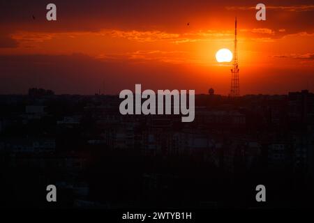 Farbenfroher rot-oranger Sonnenuntergang mit der Sonnenscheibe und der Silhouette eines Turms und Häuser in der Stadt. Stadtbild. Feuriger Sonnenuntergang über der Stadt. Stockfoto