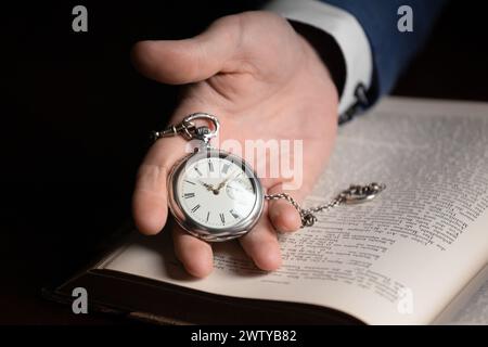 Eine Hand hält eine Vintage Taschenuhr auf dem Hintergrund eines alten Buches. Konzept von schnell fließender Zeit, Vergangenheit, Weisheit, Erfahrung Stockfoto