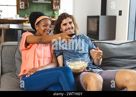 Ein vielseitiges Paar genießt einen gemütlichen Moment auf dem Sofa zu Hause, isst Popcorn und schaut fern Stockfoto