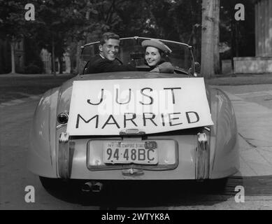 Ein junges Paar blickt von seinem Cabriolet zurück, während es wegfährt. Auf dem Heck des Autos befindet sich ein Schild mit der Aufschrift „Just Married“ Stockfoto