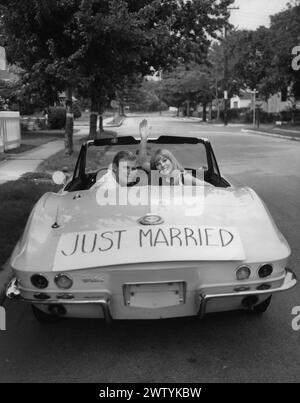 Ein junges Paar blickt von seinem Cabriolet zurück, während es wegfährt. Auf dem Heck des Autos befindet sich ein Schild mit der Aufschrift „Just Married“ Stockfoto