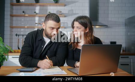 Junge Paare sitzen in der Küche und bezahlen Rechnungen mit dem Laptop, verwenden Taschenrechner Zählkosten, führen Budgetkontrolle durch, verwalten Finanzen, überprüfen überfällige Kreditzahlungen Stockfoto