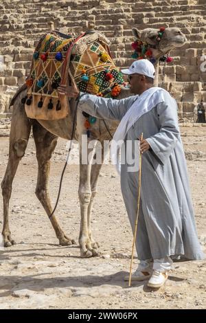 Gizeh Necropolis, Ägypten - 26. April 2022: Beduinenmann in traditionellen Kleidern, mit einem Kamel, das mit spezifischen Stickereien verziert ist, vor t Stockfoto