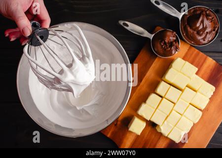 Hand hält einen Ständer Mixer Schneebesen-Aufsatz bedeckt mit Meringue: Frauenhand hält eine mit Schweizer Meringue bedeckte Drahtpeitsche über einer Metallrührschüssel Stockfoto