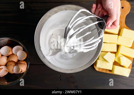 Hand hält einen Ständer Mixer Schneebesen-Aufsatz bedeckt mit Meringue: Frauenhand hält eine mit Schweizer Meringue bedeckte Drahtpeitsche über einer Metallrührschüssel Stockfoto