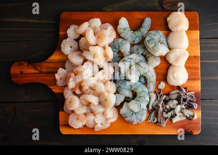 Rohe Meeresfrüchte auf einem hölzernen Schneidebrett: Ungekochte geschälte und entschäumte Garnelen, in Scheiben geschnittene Tintenfische und Jakobsmuscheln auf einem Holzschneidebrett Stockfoto