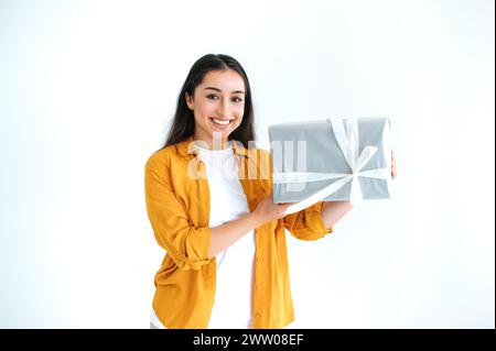 Fröhliche glückliche Brünette inder- oder arabianerin in lässigem orangenem Hemd, steht auf isoliertem weißem Hintergrund, hält große Geschenkbox, blickt in die Kamera und lächelt freundlich. Geschenke und Überraschungen Stockfoto