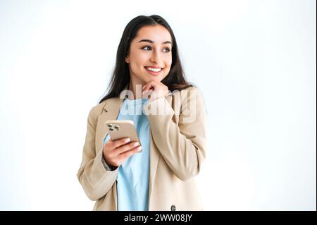 Nachdenkliche hübsche indische oder arabische Frau in elegantem Anzug, die ihr Handy für Nachrichten mit Freunden in sozialen Medien benutzt, auf isoliertem weißem Hintergrund steht, nachdenklich wegschaut, denkt, lächelt Stockfoto