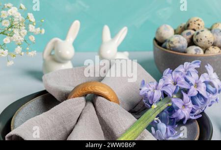 Nahaufnahme Ostertisch servieren. Graue Serviette mit Hyazinthen auf grauen Tellern, Wachteleier, Keramikhasen. Türkisfarbener Hintergrund. Horizontales Format. Stockfoto