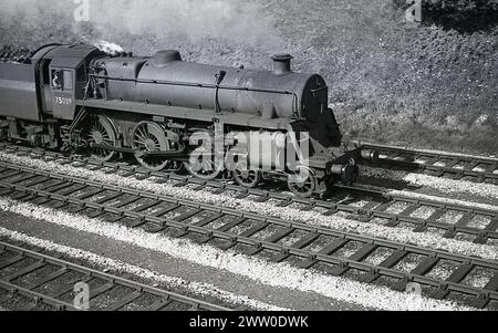 1950er Jahre, historische Dampflokomotive der Briitsh Railways, 75039 auf der Schiene, England, Großbritannien, Zugführer in der Kabine. Die im Werk Swindon gebaute und 1951 für die neuen Briitsh Railways eingeführte Lokomotive 75039 war eine BR Standard Class 4, eine kleinere, leichtere Version der Standard Class 5 7300. Sie wurde 1953 erbaut und wurde zunächst der Bletchley-Linie (1E) zugeordnet, die bis 1967 in Betrieb war, bevor sie im folgenden Jahr verschrottet wurde. Stockfoto