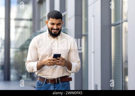 Ein lächelnder junger muslimischer Mann läuft in einem Hemd auf der Straße in der Nähe von Geschäftszentren und schaut auf den Smartphone-Bildschirm, schreibt, liest Nachrichten. Stockfoto
