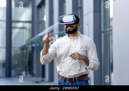 Ein junger Mann mit Bart steht vor einem modernen Gebäude, trägt ein Virtual-Reality-Headset und bewegt mit den Händen Gesten, als ob er mit der virtuellen Umgebung interagieren würde. Das Bild weckt ein Gefühl von Eintauchen und technologischem Fortschritt. Stockfoto