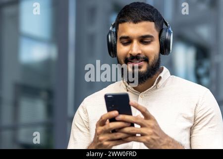 Ein fröhlicher junger Mann mit Bart sieht man draußen mit seinem Smartphone und hört Musik über Over-Ear-Kopfhörer, die eine Atmosphäre der Entspannung und des Genusses in einer urbanen Umgebung ausstrahlt. Stockfoto