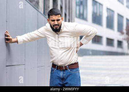 Ein müder muslimischer Mann steht draußen und lehnt sich an die Wand des Gebäudes, hält seine Hand hinter dem Rücken, grimmig und spürt starke Schmerzen im unteren Rücken. Stockfoto