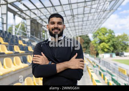 Ein junger Mann lächelt selbstbewusst mit überkreuzten Armen, steht in einem leeren Außenstadion mit gelben Sitzen und klarem Himmel. Stockfoto