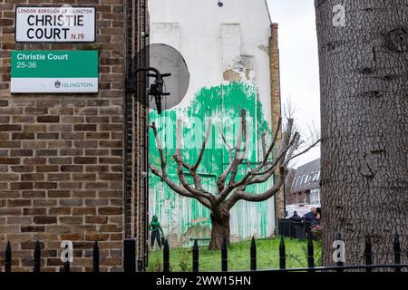 London, Großbritannien. März 2024. Neben einem neuen Kunstwerk des weltbekannten Straßenkünstlers Banksy, das am Vortag auf der Seite eines Wohngebäudes hinter einem Baum im Garten eines Wohnblocks im Finsbury Park erschien, ist ein Schild des Islington Council Social Housing abgebildet. Das Kunstwerk, von dem der Straßenkünstler behauptet wurde, hat die Form einer grünen Farbe, die auf eine Wand hinter einem bestückten Kirschbaum gesprüht wird, um von vorne gesehen wie Laub zu erscheinen, zusammen mit einer Schablone einer Frau, die einen Hochdruckreiniger hält. Der verwendete Grünton scheint mit dem verwendeten Farbton zu übereinstimmen Stockfoto