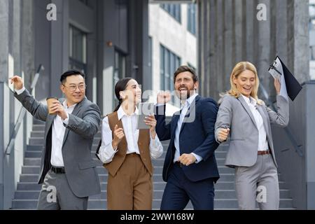 Eine vielfältige Gruppe begeisterter Geschäftsleute feiert einen Sieg oder Erfolg mit Faustpumpen und fröhlichen Ausdrücken vor einem modernen Bürogebäude. Stockfoto