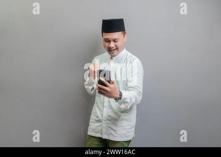 Asiatischer muslimischer Mann, der ein koko-Shirt und Peki in den Farben des Fastenmonats trägt, seine Faust zusammenklammert und sein Smartphone hält, während er auf Smartp schaut Stockfoto
