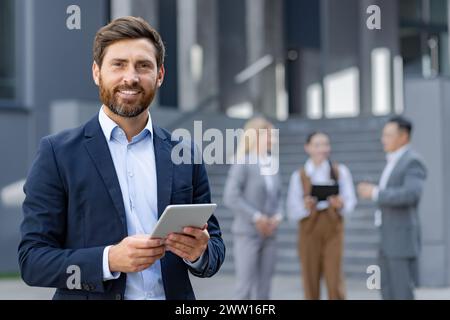 Lächelnder Geschäftsmann mit einem Tablet mit verschwommenen Kollegen im Hintergrund, der Führungsqualitäten und Technologieintegration in einer modernen Unternehmensumgebung veranschaulicht. Stockfoto