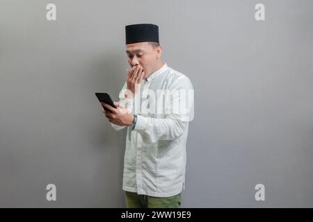 Überraschter asiatischer muslimischer Mann, der Smartphone-Handy für Anrufe, soziale Medien, mobile Anwendung online benutzte, ein koko-Shirt und Peki mit Farben trug Stockfoto