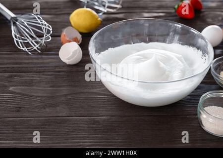 Schüssel mit geschlagenem Eiweiß zum Backen, mit einem Schneebesen, frischer Zitrone und Eierschalen auf einer hölzernen Arbeitsplatte. Stockfoto